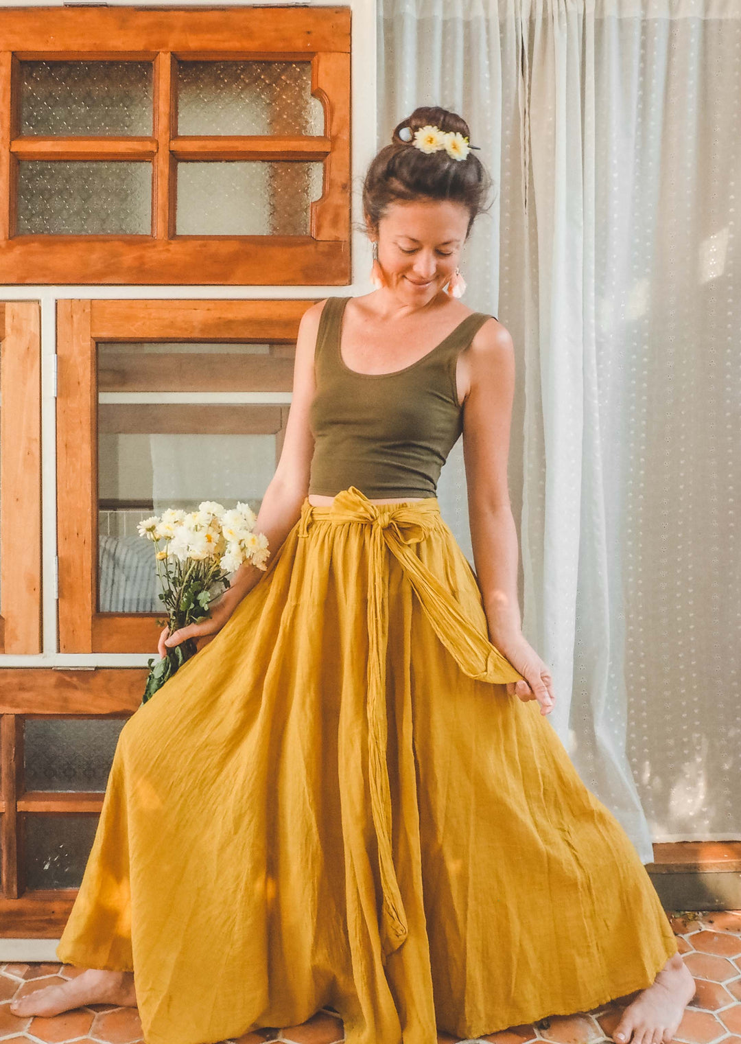 Woman has on olive green tank top and gold-color maxi skirt. She is holding daisies in right hand.