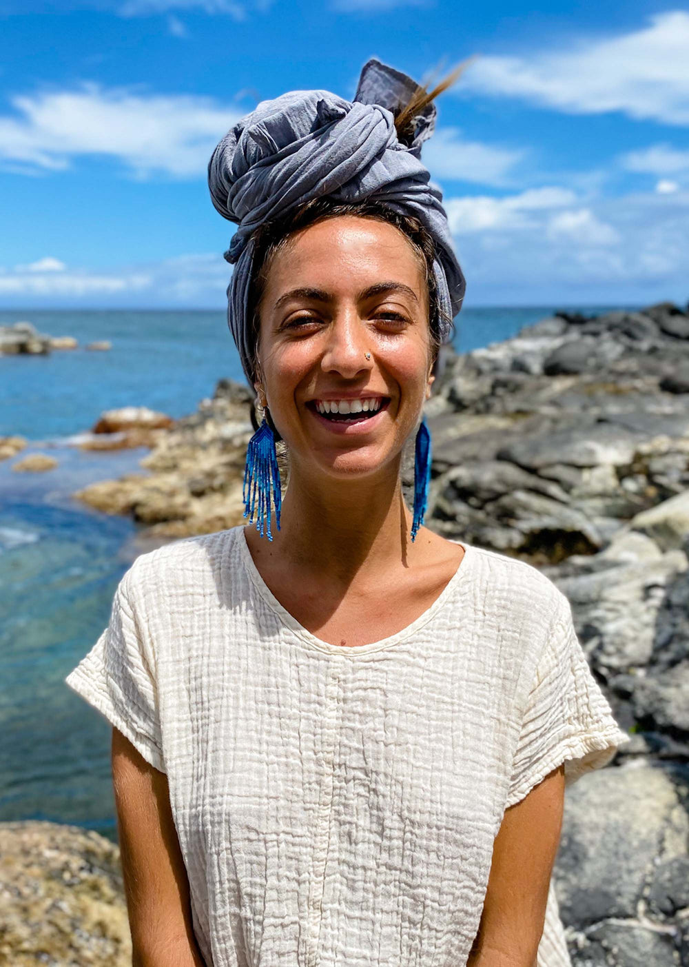 Model stands by ocean with hair tied up in long blue scarf.