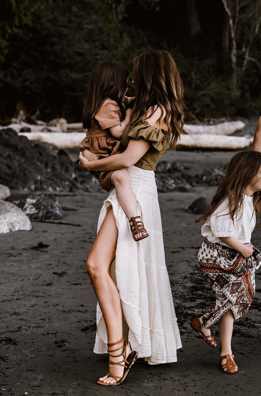 Woman wearing long white skirt and brown sandals holds little girl at waist.