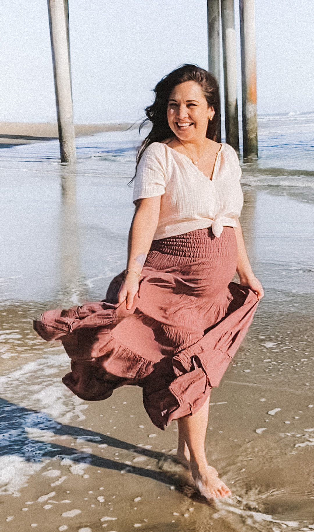 Maternity clothing model wears white top paired with long, stretchy, flowing skirt while walking in the water at the beach.