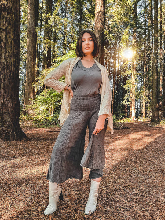 Female model poses among trees wearing pants with elastic waist, tank top, cardigan and boots.