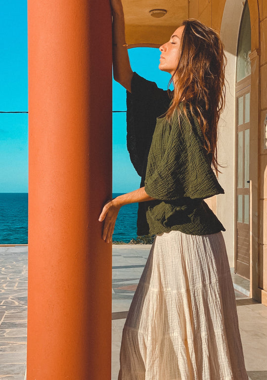 Woman stands at pillar wearing flowing green top and white skirt. 